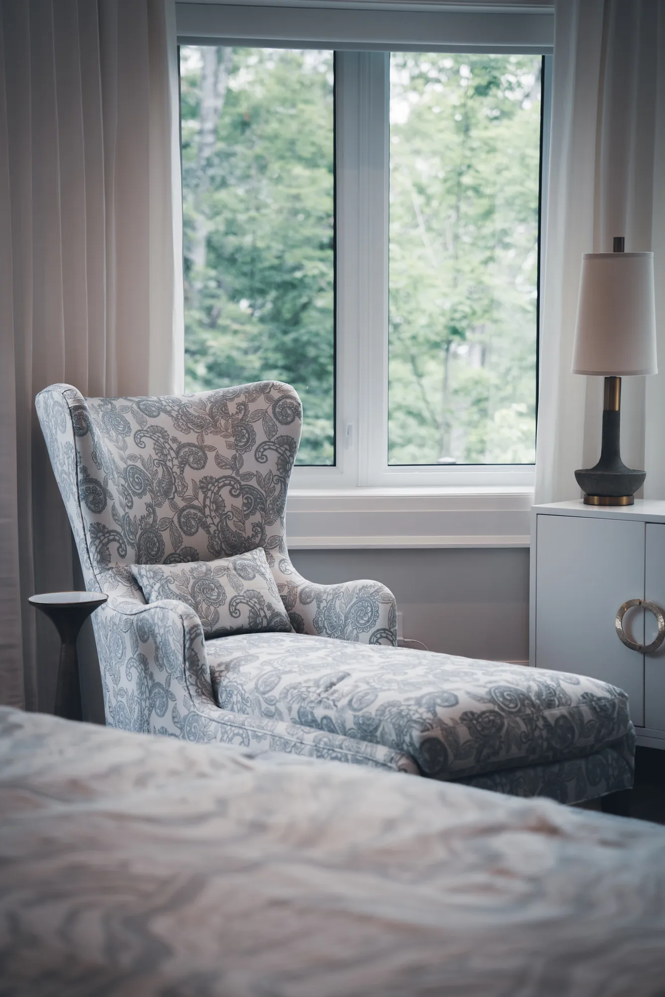 A paisley wingback chair with a long seat by a bright window, beside a white side cabinet and lamp