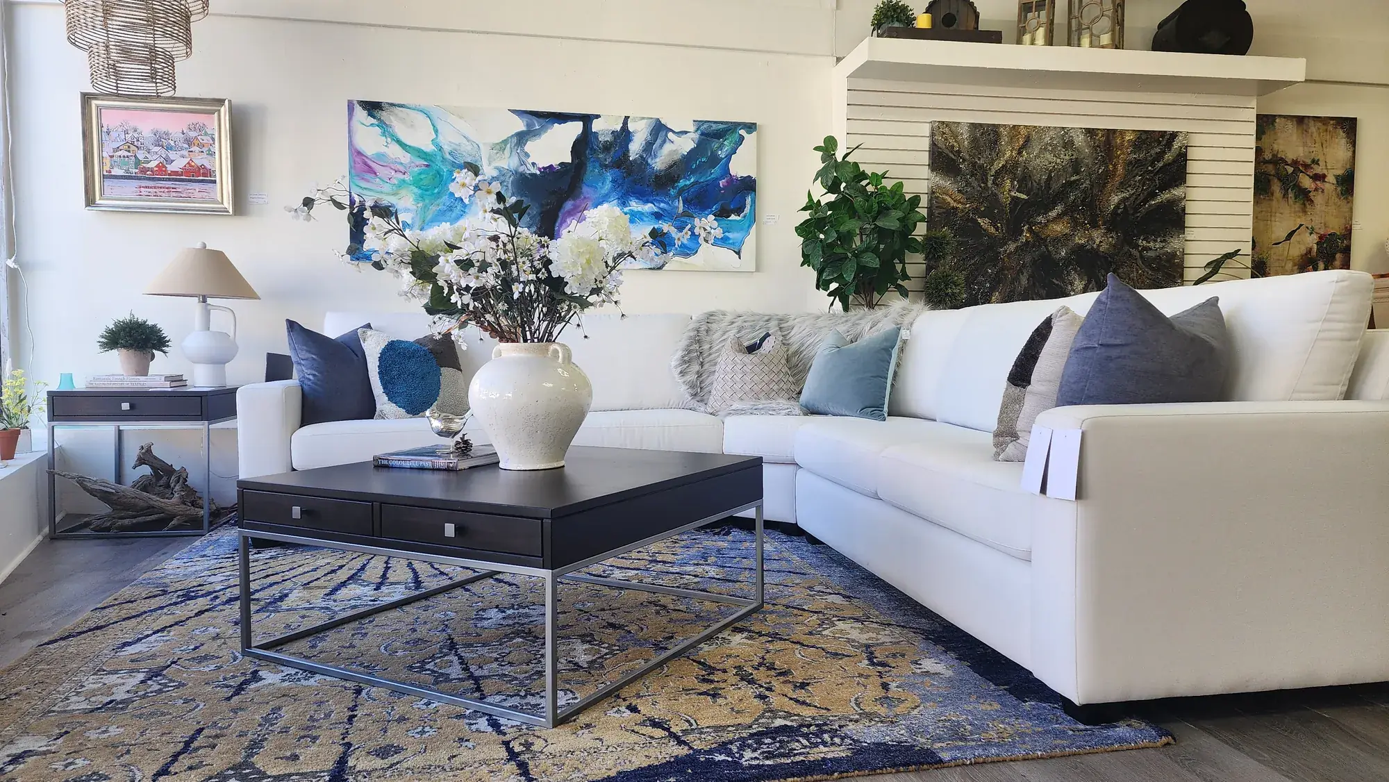 A white leather sectional and a dark coffee table on a blue patterned rug in the showroom, with abstract paintings above